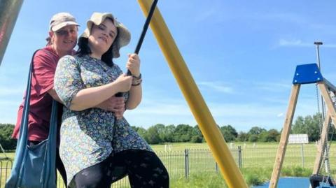 A young woman is on a swing. She is wearing a sun hat and looks happy. A woman is standing behind her, smiling. It's a sunny day and both women look happy.