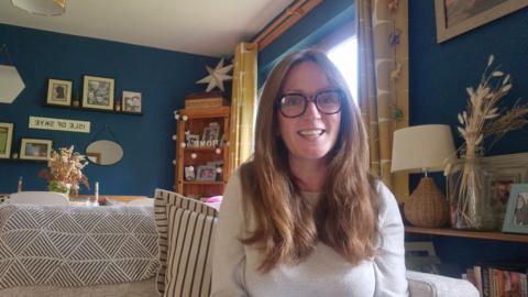 Kathryn NicAoidh, na suidhe air beulaibh chamera agus gàire air a h-aodann.