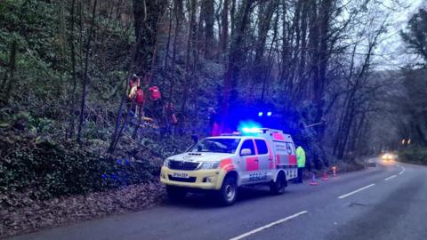 A roadside cave rescue operation under way, with a specialist vehicle on blue lights next to a wooded bank