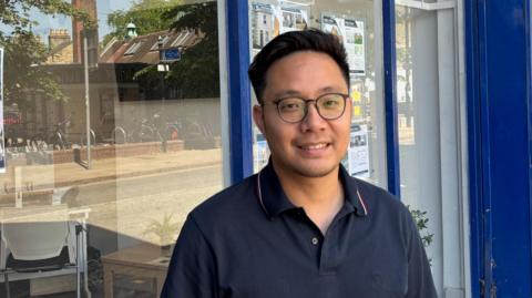 Ray Yu, a young man with short, dark hair, glasses and wearing a blue polo shirt, standing outside a shop.