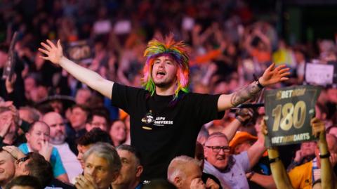 A crowd of people watching a darts contest at Glasgow's Hydro. A man in the middle of the crowd is on his feet, wearing a multi-coloured wig and a black t-shirt. His arms are out-stretched and his mouth open wide. He's enjoying himself. 