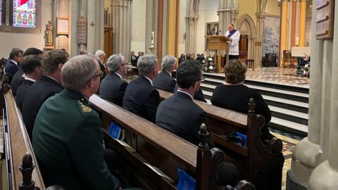 People sit on church benches as the Canon Dominic Golding gives a speech from the altar. There is a stained glass window in the background and statues and drawings of religious moments on the walls. 