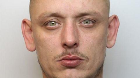 A police mugshot of a man with looking at the camera, with a moustache and hazel eyes.