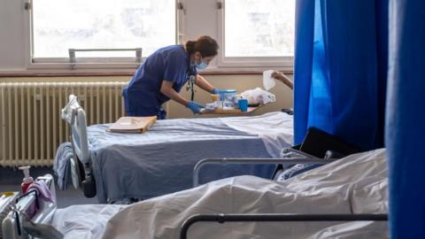 Inside a hospital ward, two patients can be seen lying in hospital beds. Their faces can't be seen as they are behind the divider curtains. A nurse wearing a face mask speaks to one of them.