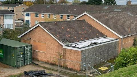 The exterior of the building with a green container next to it. It is in need of repair with missing roof tiles.