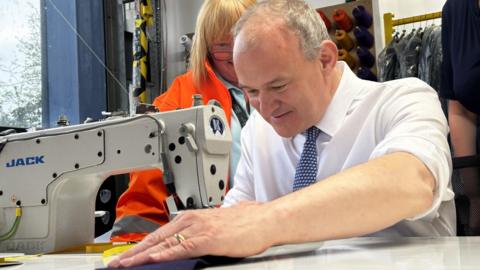 Sir Ed Davey holds his hand over a piece of material he has placed under the needle of a sewing machine in a factory. He is wearing a white shirt and tie and is smiling. A woman in an orange high-vis jacket watches on behind him. 