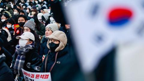 A South Korean flag waves in the foreground, with a crowd of people gathered in the background, one holding a placard that says 'Arrest Yoon Suk Yeol right now', in Seoul on 8 December