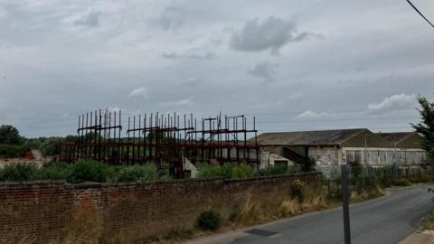 Remnants of a Victorian factory behind a brick wall. Structures pointing up to the grey sky. Buildings to the right and a road in the foreground.