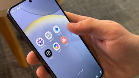A hand holding a black mobile phone with six apps on the screen: Meta AI, ChatGPT, Gemini, Grok, Copilot, Claude.