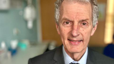 UEA vice‑chancellor Professor Maguire wearing a grey suit blue check shirt and tie looking at the camera.