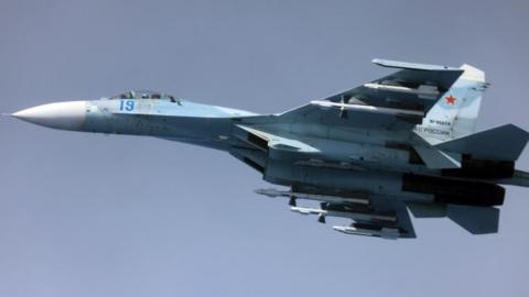 Image of a Russian Su-27, a whit and blue coloured fighter jet, airborne against a clear blue sky.