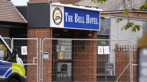 Image of the front of the Bell Hotel which is surrounded by a metal fence. A police van is parked outside. 
