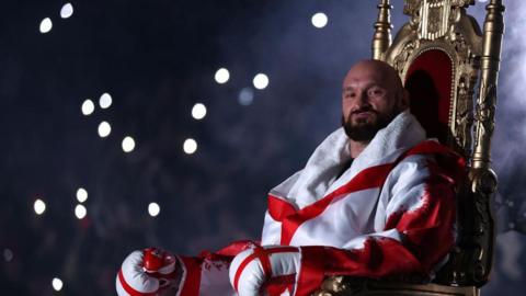 Tyson Fury smirks as he enters the ring on a throne, dressed in red and white