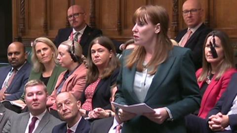 Angela Rayner standing and speaking in the House of Commons chamber, holding papers. The chamber is filled with people seated on green benches, with wood-paneled walls.