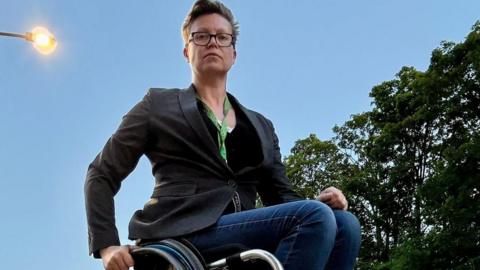Sarah Hope is photographed on a station platform in a wheelchair. A streetlight is on and there are trees in the background. She is photographed from the ground and she is looking down at the camera.