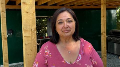 There is a woman with brown shoulder-length hair, a pink flowery blouse and a flower pendant around her neck. She is stood underneath a wooden frame which is used by the snack shack to stay dry. Behind her is the shack, which is painted dark green.