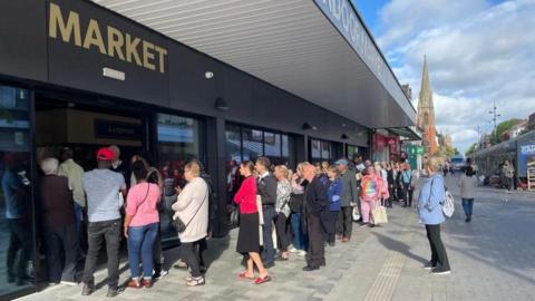People are standing in a long queue outside a large building. The street can be seen in the background and there are people walking by and shops in the distance.