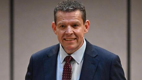 Rhun ap Iorwerth looking into a camera, wearing a dark blue suit and a light blue shirt. He is wearing a red tie with white spots.