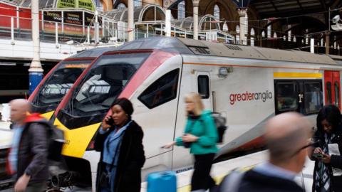 People on Liverpool Street station platform with a Greater Anglia train behind them. Some a on mobile phones others are walking with luggage.