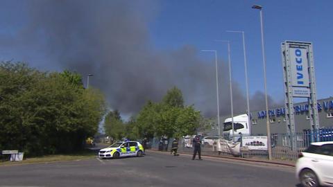 A large black smoke plume can be seen blowing over an IVECO garage with a parked lorry behind a large fence. A police car is parked across the street and a firefighter is walking in the direction of the smoke.