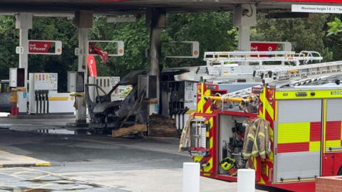 A van which has been damaged by a fire with a fire engine in attendance