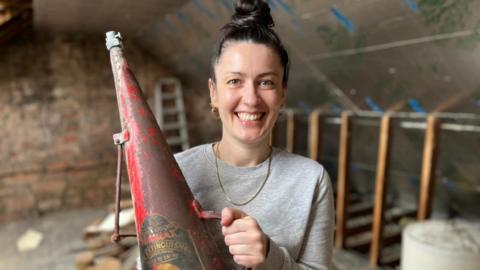 Kirsty Geddes with tied back brown hair, smiling to camera, wearing a long-sleeved grey top and holding a red cone-shaped metal fire extinguisher dated 1942