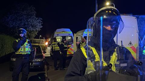 Police officers wearing riot gear stand in front of police vehicles. Several police vans  can be seen in the background. A blue light glows on the plastic shield one officer holds in front of his face 