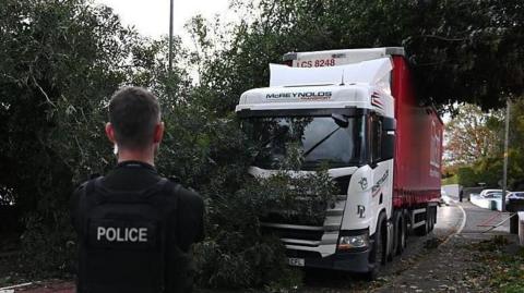 Police officer in black clothing in front of fallen tree and white and red lorry