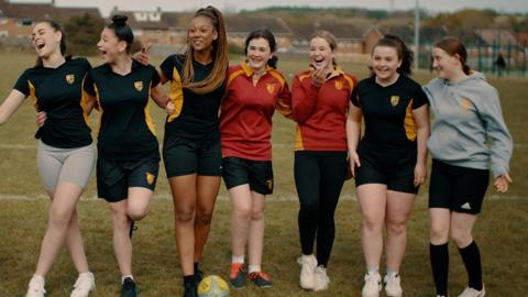 Grŵp o ferched mewn crysau ysgol chwaraeon yn chwerthin ar y cae, gyda phêl rygbi wrth eu traed.