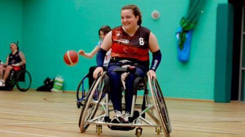 Libby wearing her wheelchair basketball team uniform. She is in her wheelchair on a basketball court and is smiling.