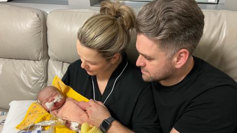 Bryony and Daniel Russo, both wearing black, sit on a cream sofa while cradling Emmy, who has tubes attached to her and is lying on a yellow blanket.