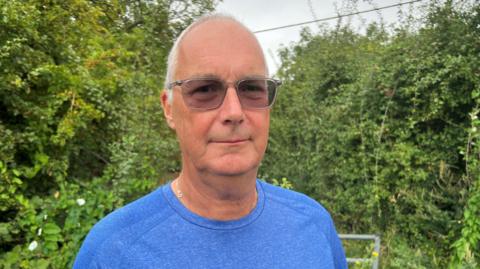 Mark Kelby - a man with thin grey hair and tinted glasses - looks straight at the camera. He is wearing a blue T-shirt and is standing in front of an area of hedge and trees.