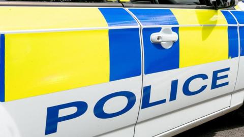 A side view of a UK police car with "police" in blue letting along the side against a largely white background, with one yellow stripe visible