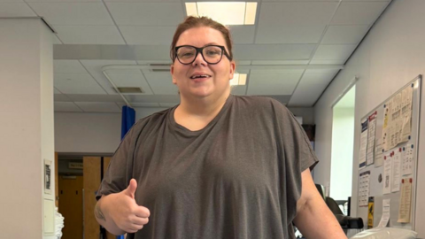 Natalia is at a physio rehab centre, stood between parallel bars to help her walk. Her left leg is in a metal cage. She makes a thumbs up pose to the camera. 