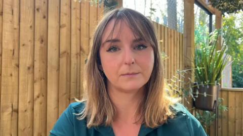 Crystal has long, brown shoulder-length hair and is wearing a green top. She is sitting outside by a wooden fence with plants behind her. The sun is shining.