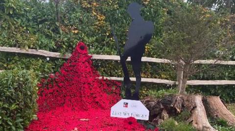 A silhouette of a soldier stands on grass next to a tree trunk, with a carpet of thousands of red knitted and crocheted poppies in front and beneath his feet