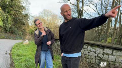 A woman with a country style jacket and jeans wearing glasses with her hair tied up is pointing up the road. Next to her is a man in a black sweatshirt and black jeans he is also pointing up the road concerned about poor driving