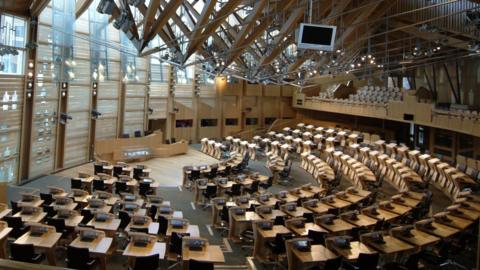 An empty Holyrood chamber - several rows of pine desks are arranged in a semi-circle facing the presiding officer's lectern at the front, while soft light streams in from above.