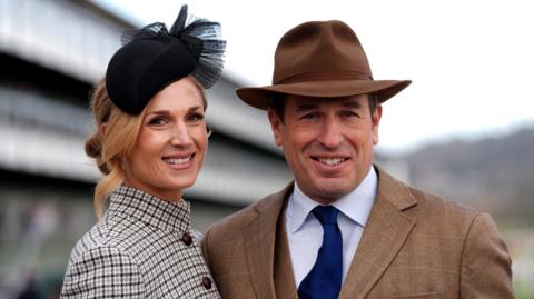 Peter Phillips and Harriet Sperling posing for a picture at a formal event. He is wearing a brown hat and matching tweed jacket with a dark blue tie. She is wearing a houndstooth-patterned blazer buttoned up to the top, and a black fascinator with gathered mesh detail. They are standing close together and smiling at the camera on a cloudy day.