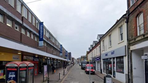 A google street view image showing a high street with shops along the sides, vans parked on the edge of the road as well as shoppers and phone boxes.