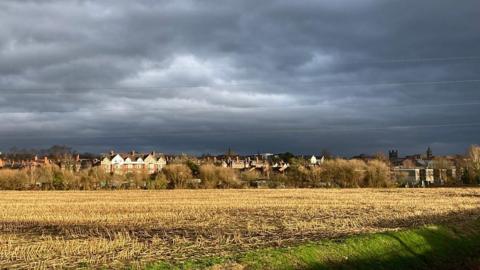 Dark grey clouds sit over a row of houses with a field in the foreground reflecting the sun