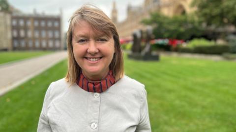 Sarah Gibson, the MP for Chippenham, stands on College Green in London, smiling at the camera. She is wearing a beige jacket and red scarf.