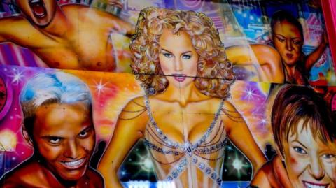 A highly colourful airbrushed artwork featuring a a blonde woman wearing a bejewelled leotard, she is surrounded by four other figures against a kaleidoscopic background.