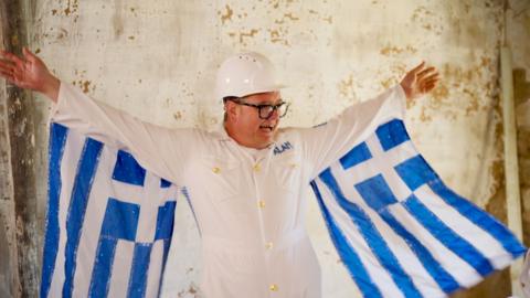 Alan Carr in a white hard hat and jump suit with Greek flags draped like wings from his arms
