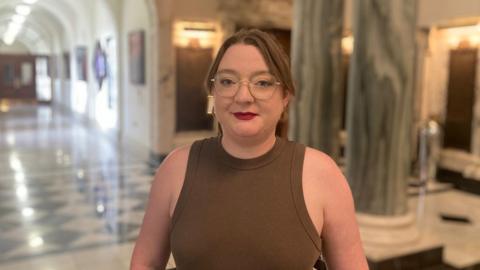 A woman in a brown vest is looking at the camera. She wears glasses and red lipstick. Behind her are marble pillars and an arched hallway. 