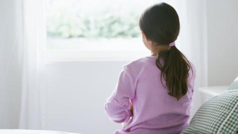 Young girl sitting on edge of bed looking out a window