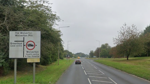 A road with grass verges and trees on either side. There is a sign on the left which points towards Old Wolverton and Wolverton Mill.