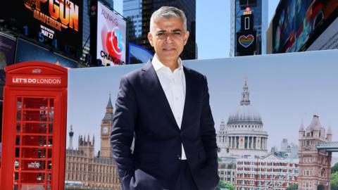 Dressed in a navy suit and white shirt the Mayor of London Sadiq Khan  poses at Times Square in front of a billboard showing London landmarks.