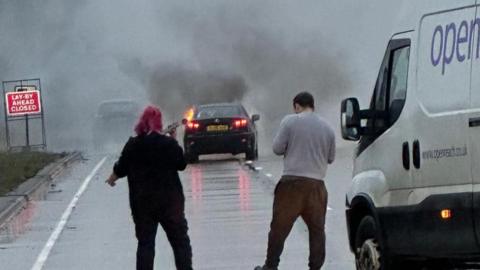 A woman with pink hair holds a phone to her face and stands next to a man next to a white van in a rainy carriageway as they look at plumes of grey smoke rising from a back car.