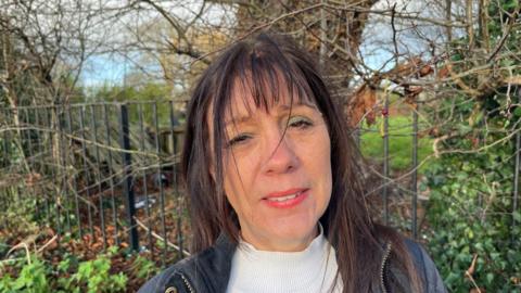 Hazel Harwin, 61, has black hair over her shoulders with a fringe across her forehead, she is wearing a black leather type jacket with white jumper underneath. Behind her is 9m (30ft) tall horse chestnut tree she is planning to sit in on Christmas day to try and gain more signatures for a petition she started to save it and others along Barkers Lane, where she lives.